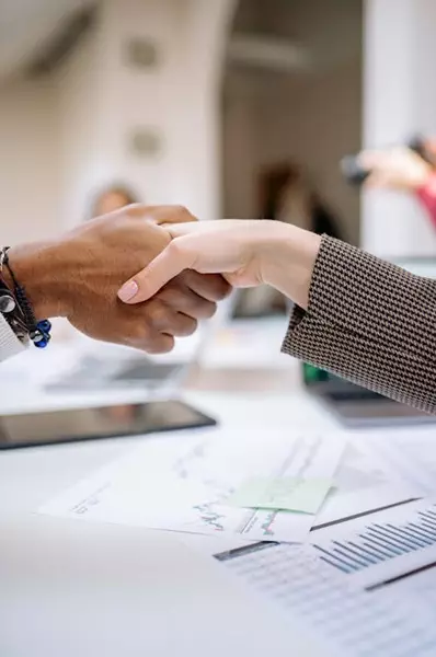 Two people shaking hands over financial documents, symbolizing an arrangement to deal in financial services.