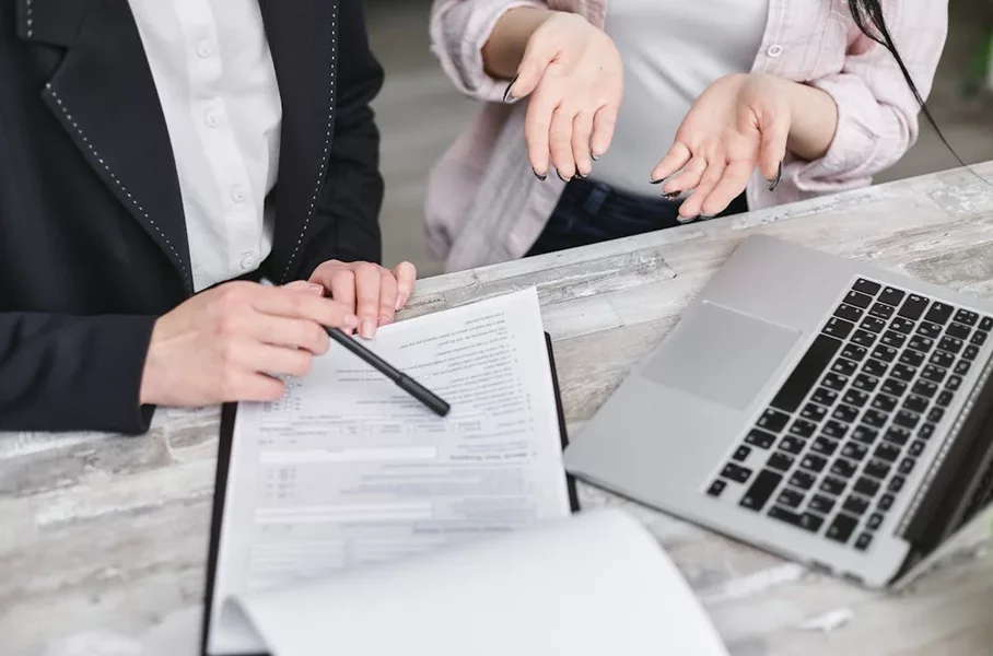 Two people discuss a financial product deal with paperwork and a laptop, focusing on an AFS licence.