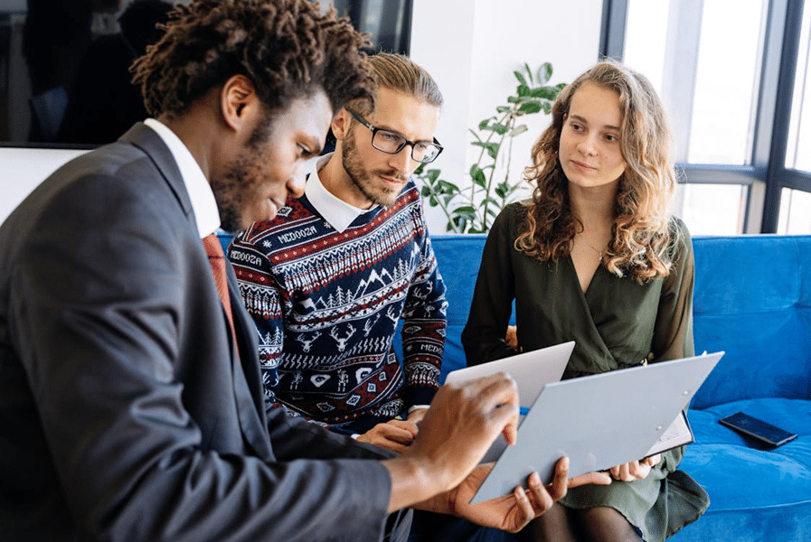 Three people discussing financial product advice, looking at documents and laptops in a modern office setting.