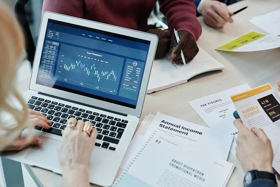 Business meeting with a laptop showing graphs, annual income statement, and financial data papers on a table.