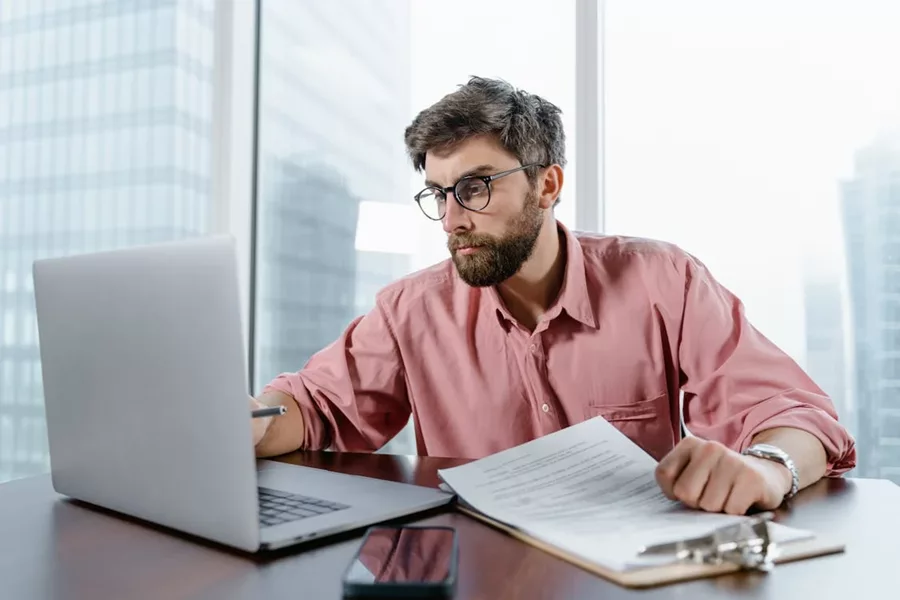 Man in glasses reviewing documents on a laptop, pondering how long the AFSL application process will take.